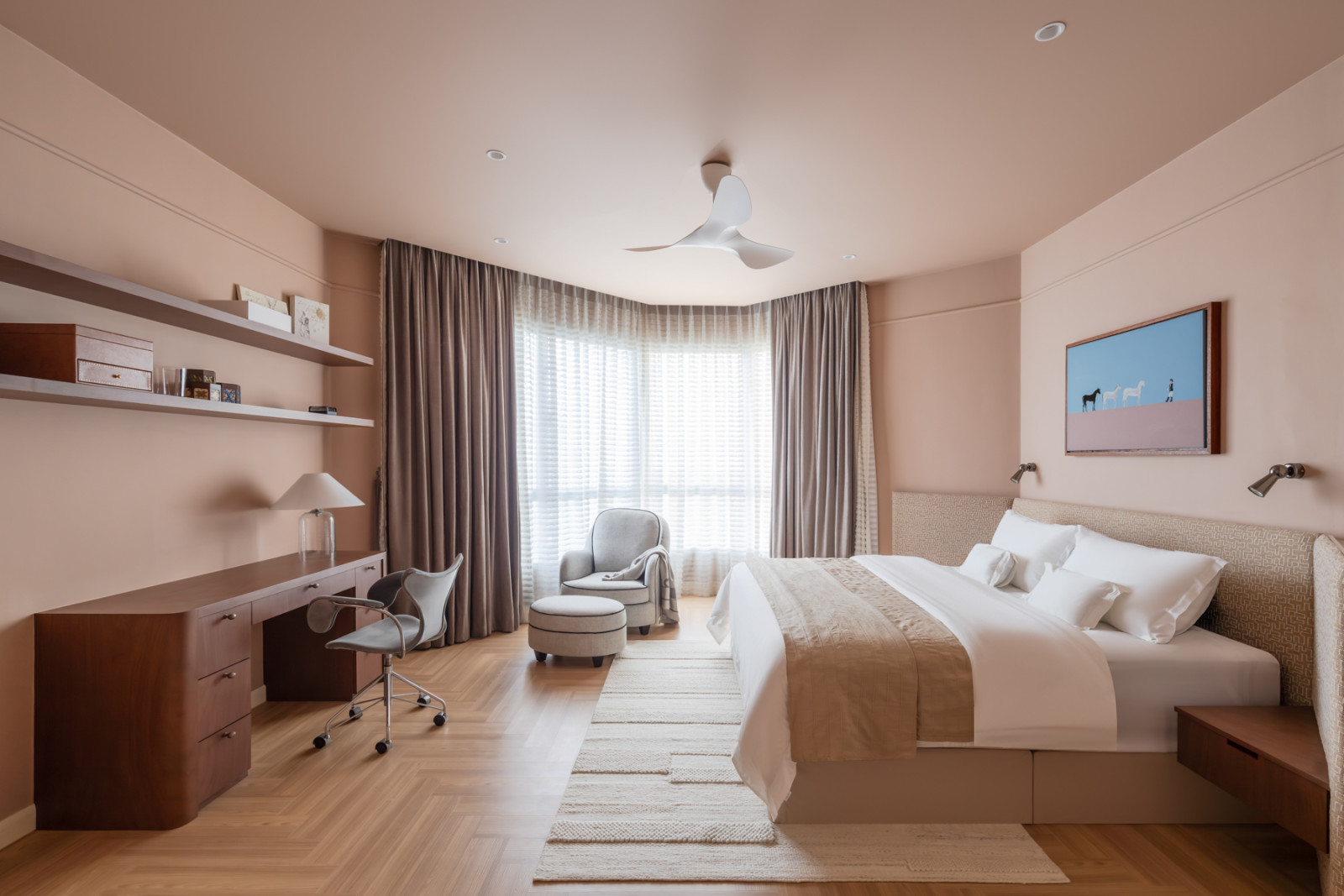 The master bedroom in full view, showing the herringbone timber floor, walnut desk, globe-base table lamp, and a small, rounded armchair and ottoman by the curtained window