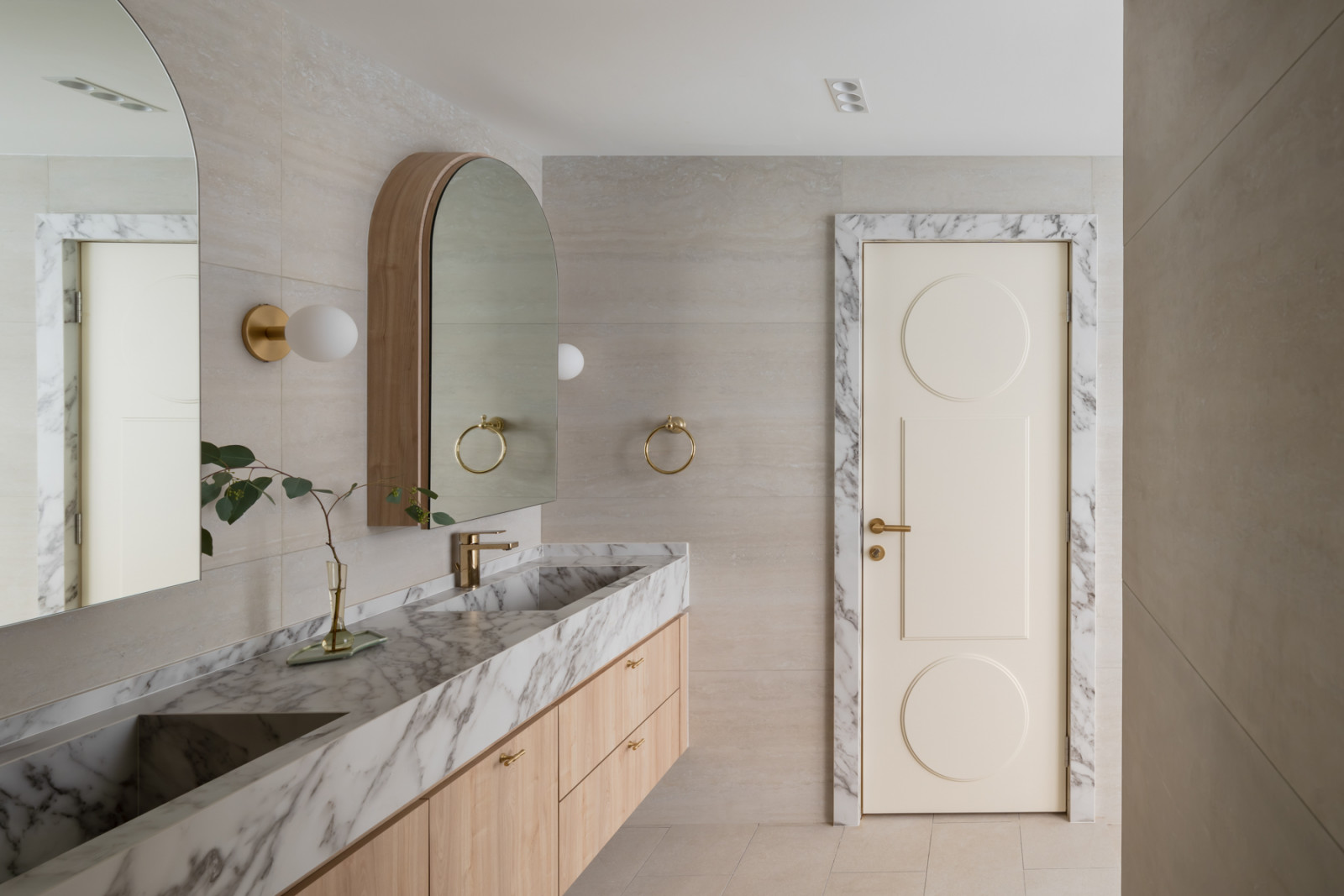 The master bathroom vanity, with a Carrara marble-edged double sink set into a light timber cabinet, an arched wood-framed mirror, and a door with circular relief detailing