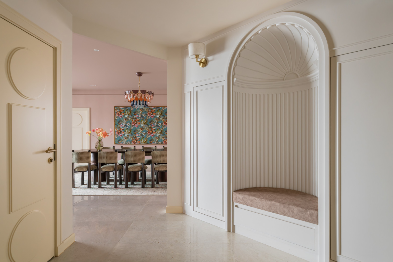 The entrance foyer, with a built-in arched nook in white plaster featuring fluted panelling and a cushioned bench, with a view through to the pink dining room beyond