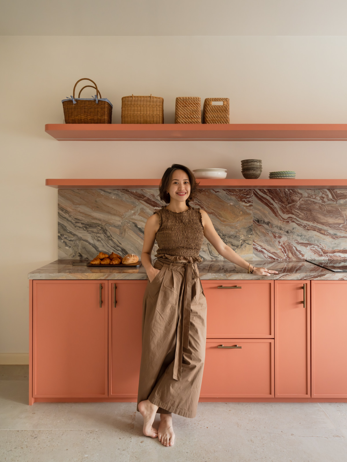Designer Shen Yee Choong in the kitchen, which features terracotta-pink cabinetry, open shelving lined with rattan baskets, and a richly veined marble backsplash