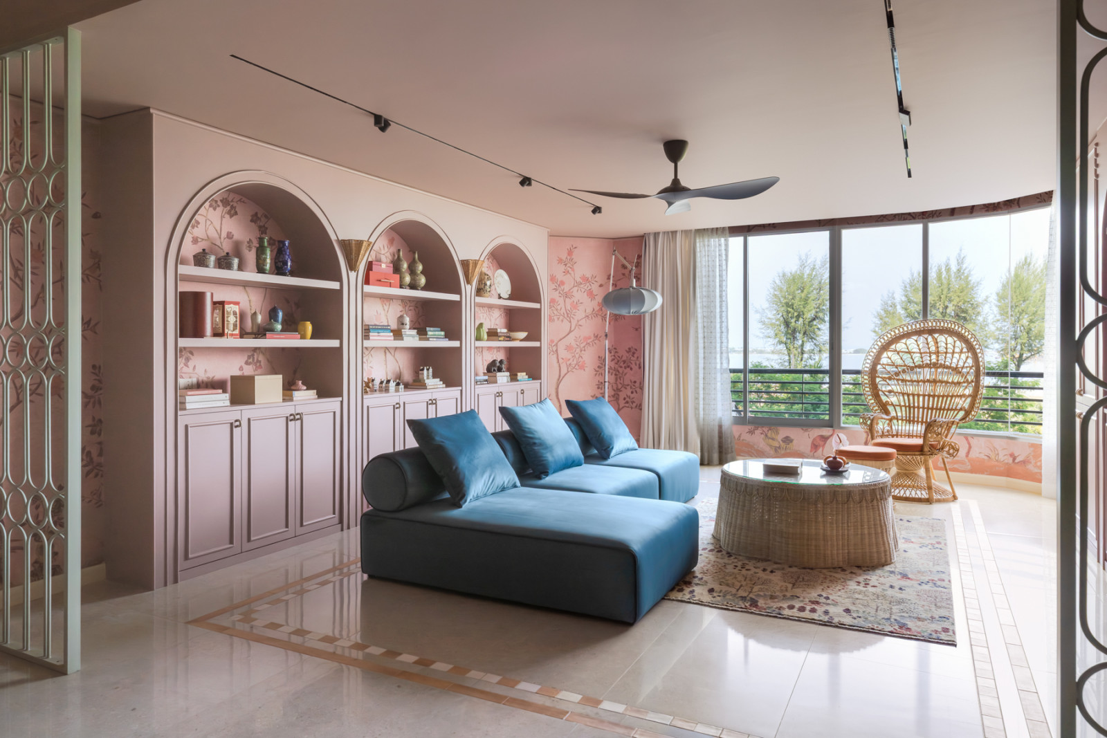 The living room in full, with the blue velvet sofa, chinoiserie wallpaper wall, arched pink shelving, rattan peacock chair, and floor-to-ceiling windows looking out to the sea and tree canopy