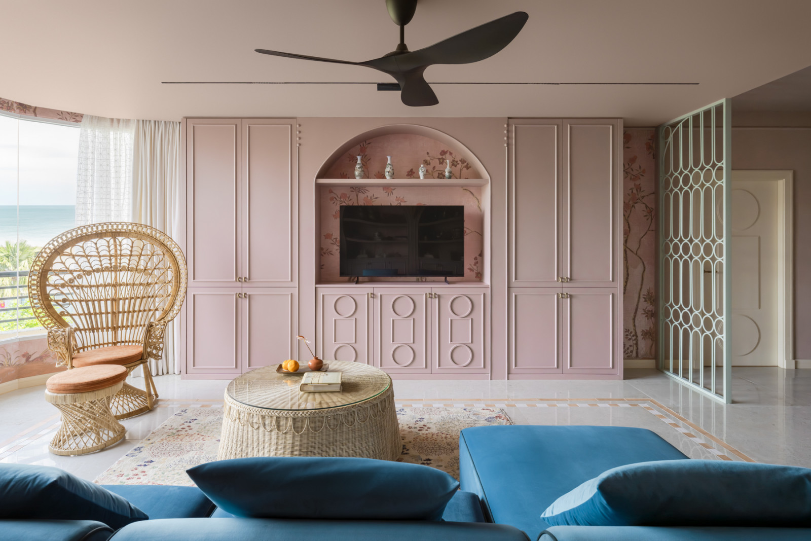 The living room faces the television wall, with pink arched built-in cabinetry featuring circular relief detailing, a rattan peacock chair, a woven rattan coffee table, and a sea view through the full-height windows