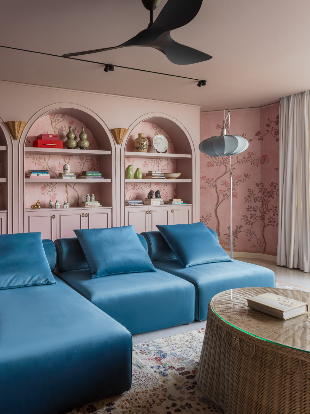 The living room, with a blue velvet modular sofa facing the pink arched shelving units lined with the owners' collected objects, books, and ceramics, set against chinoiserie wallpaper