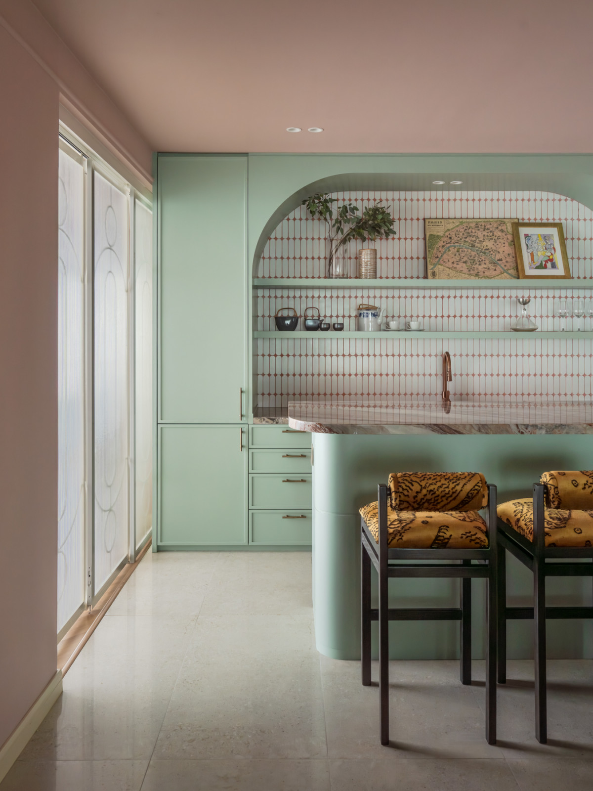 The bar, with mint-green cabinetry, a rounded island base, patterned red-and-white tile backsplash set within a plaster arch, and two bar stools upholstered in a tiger-print fabric