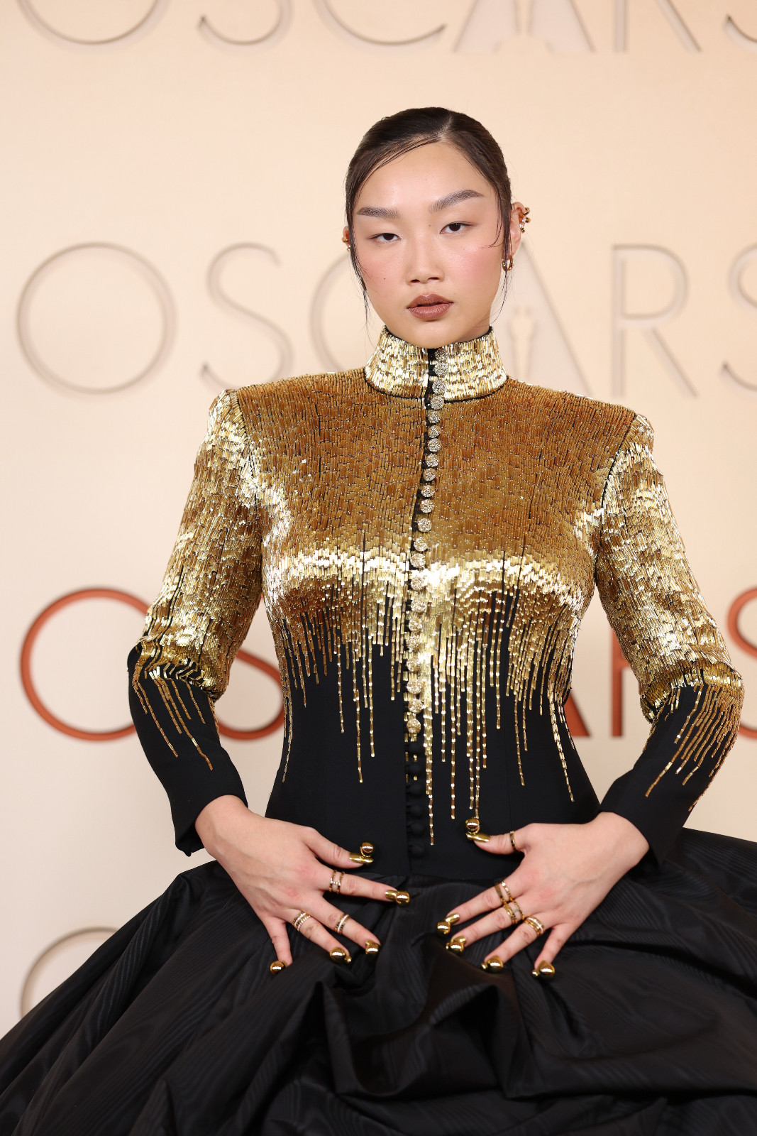Audrey Nuna brought a graphic edge in a gold‑embellished Thom Browne jacket and voluminous black skirt, paired with sleek hair, neutral makeup and sculptural gold nails (Photo: Myung J. Chun / Los Angeles Times via Getty Images)