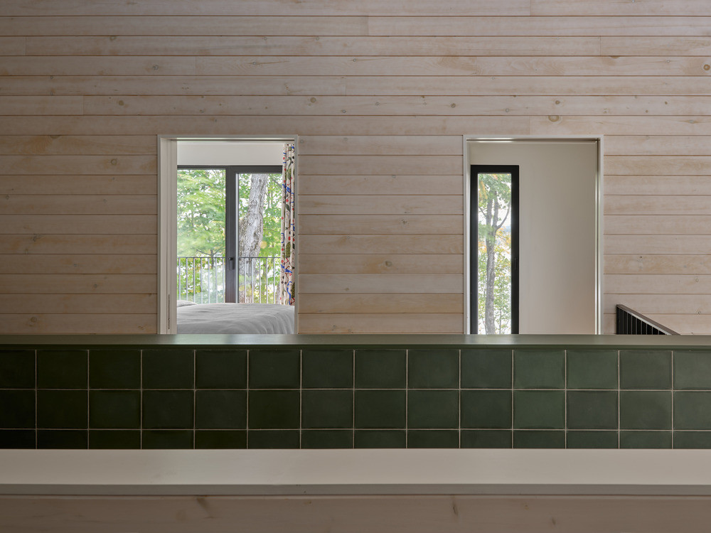 A corridor on the upper ground level, with whitewashed knotty pine walls and a green-tiled half-wall overlooking the bedroom beyond