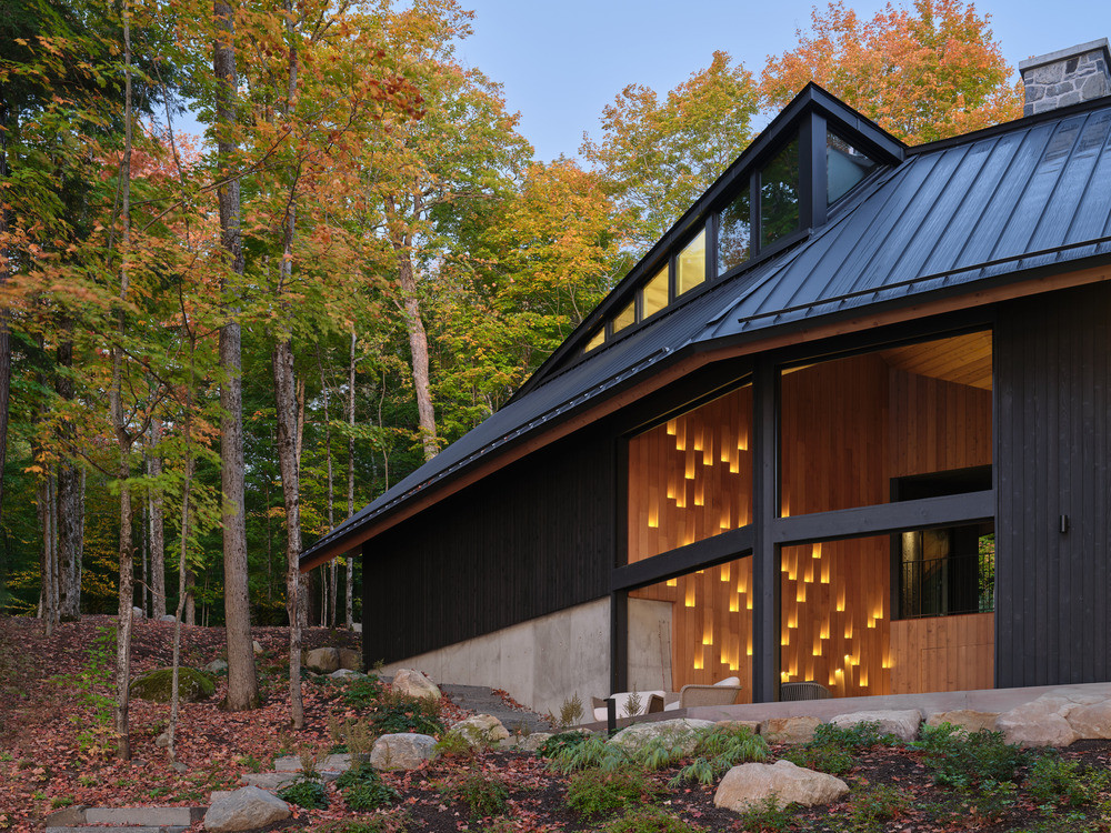 The lake-facing elevation at dusk, showing the junction between the two cedar-clad volumes and Victoria Fard’s lit cedar wall pattern visible through the glazing of the Muskoka Room