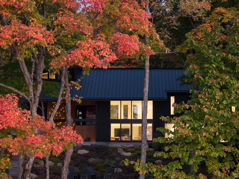 The lake-facing elevation at dusk, seen through the autumn maple grove with the clerestory windows and lower-level glazing is lit from within