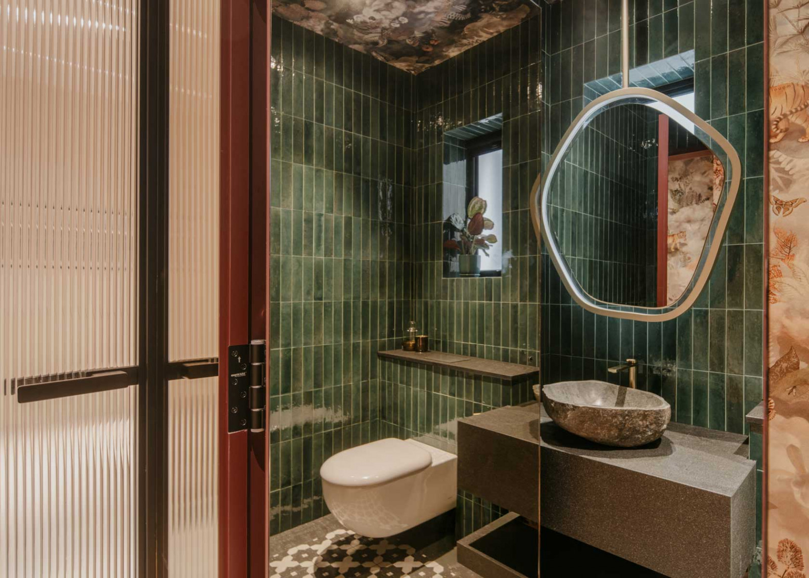 Green subway tiles, a burgundy door and an amoeba-shaped suspended mirror create a playful atmosphere in the guest bathroom