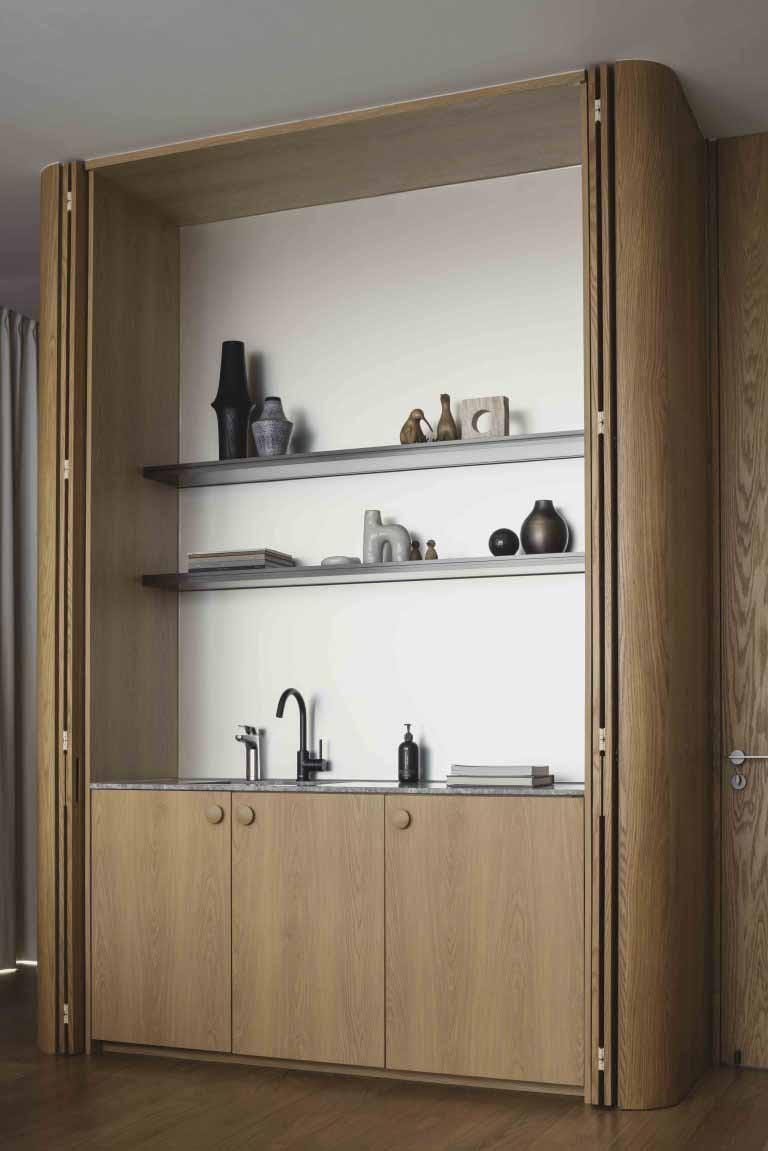 A built-in timber cabinet integrates a compact sink and open shelving within a concealed niche