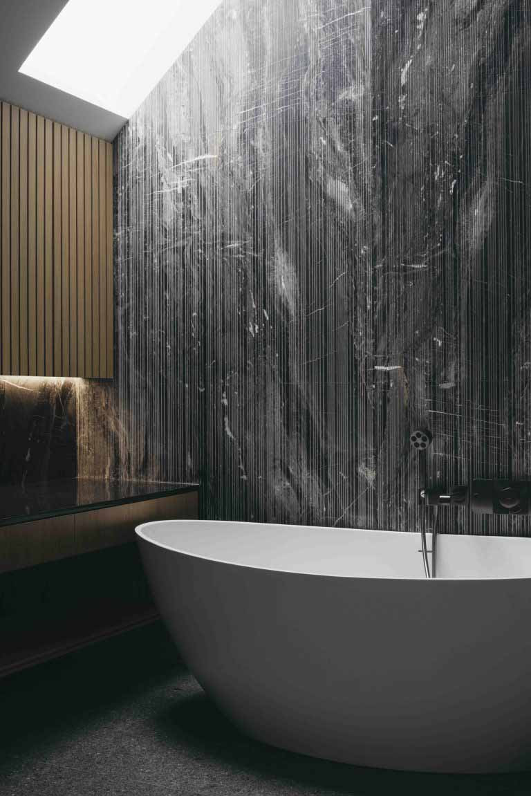 A freestanding bathtub sits against a full-height stone wall, lit by a skylight above