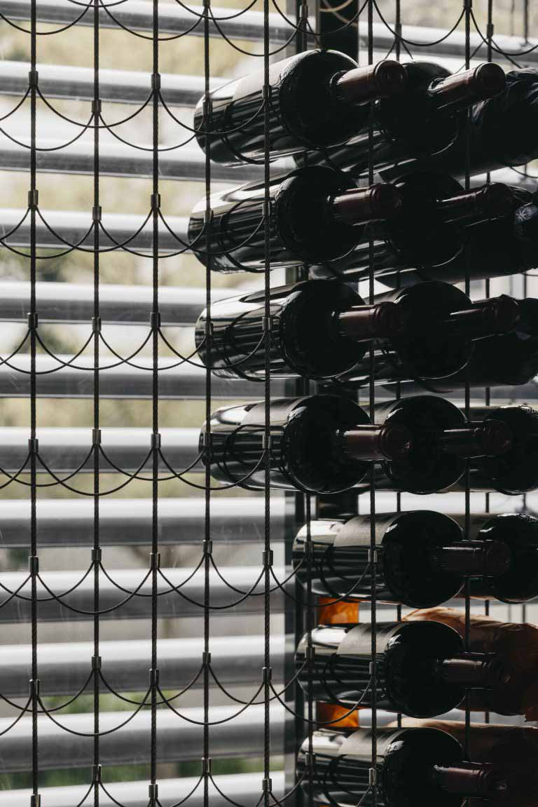 A suspended wine storage system sits in front of a screened façade, filtering daylight