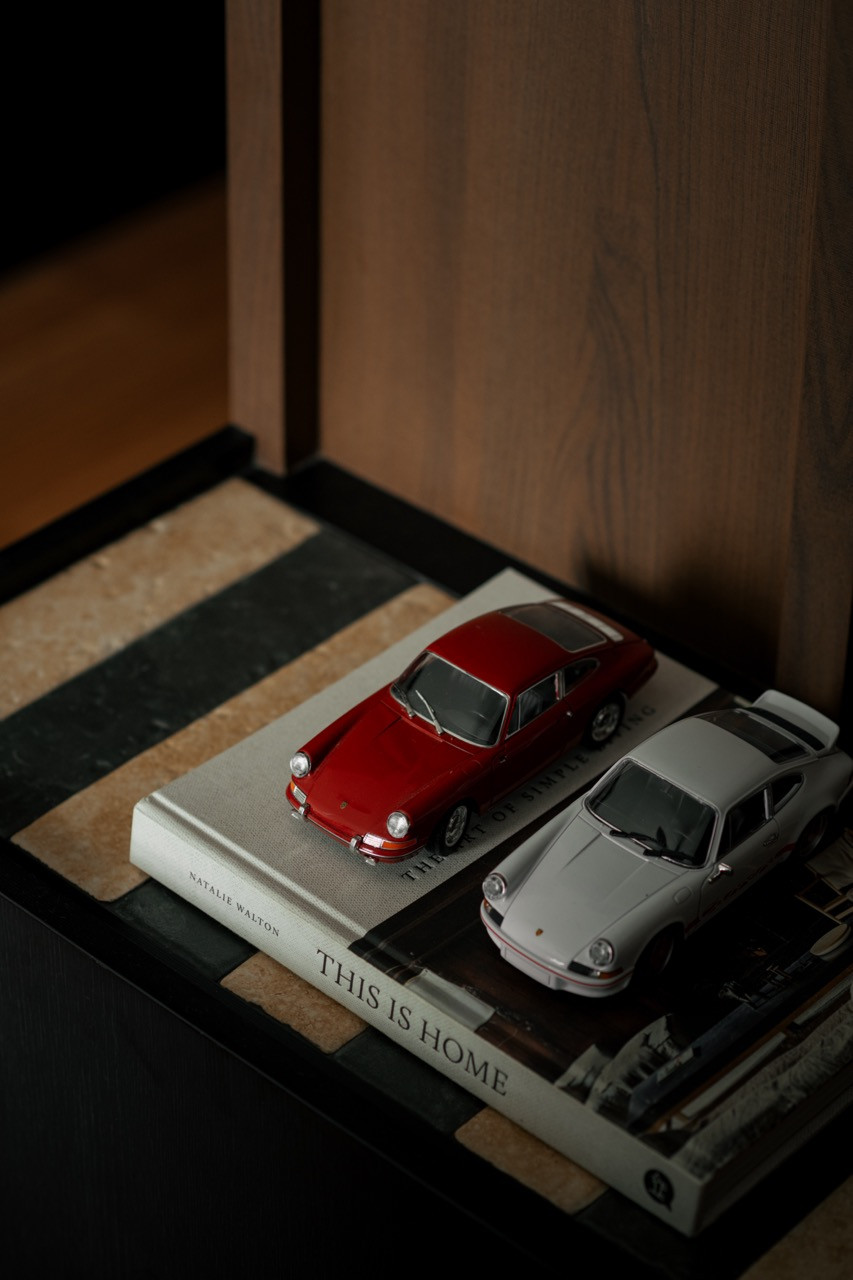 Two Porsche scale models on a copy of Natalie Walton's This Is Home, placed on a dark tray within the bedroom shelving