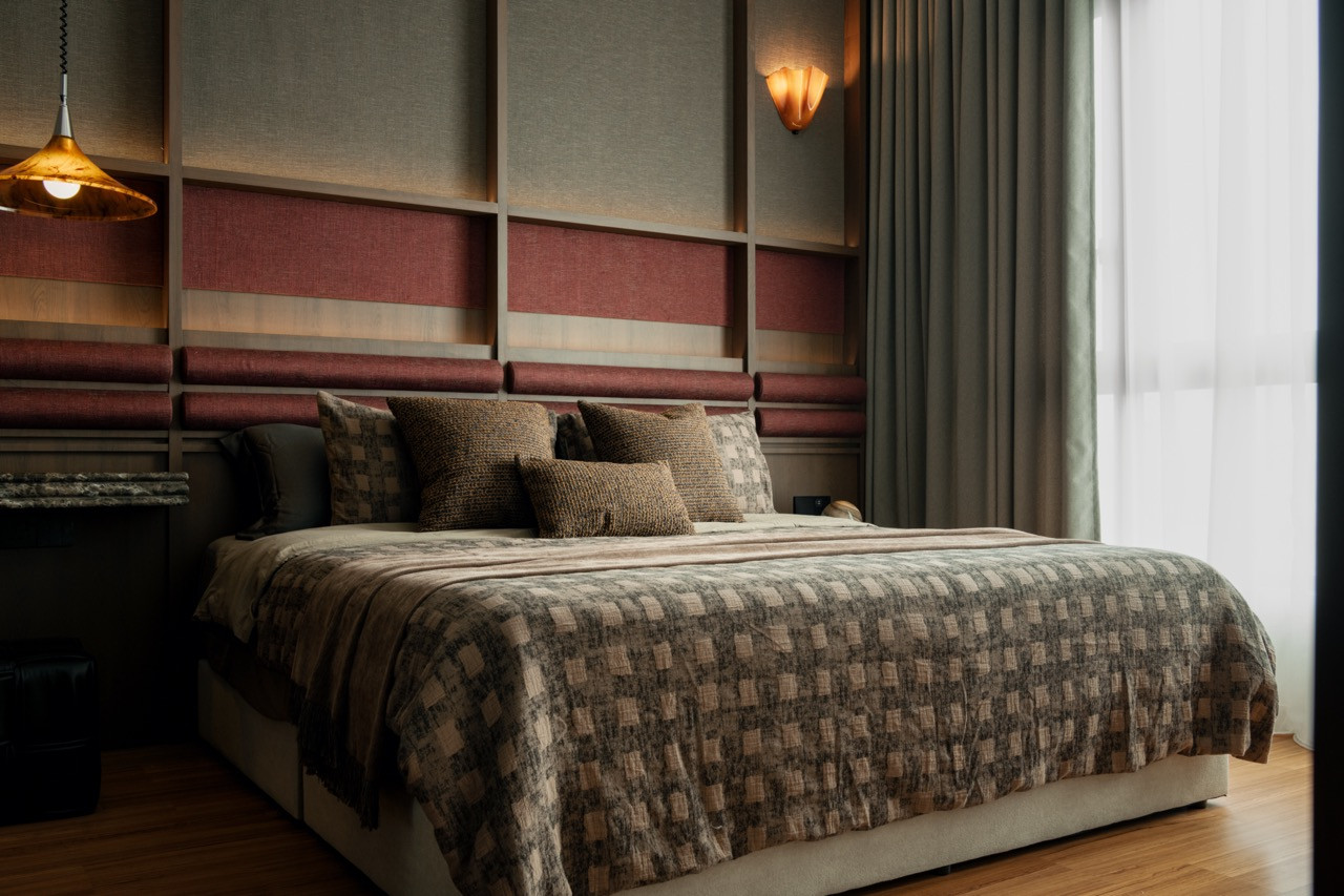 The bedroom seen from the doorway, with the full fabric-panelled headboard wall and its banded treatment of grey and crimson, curtained window and copper pendant.
