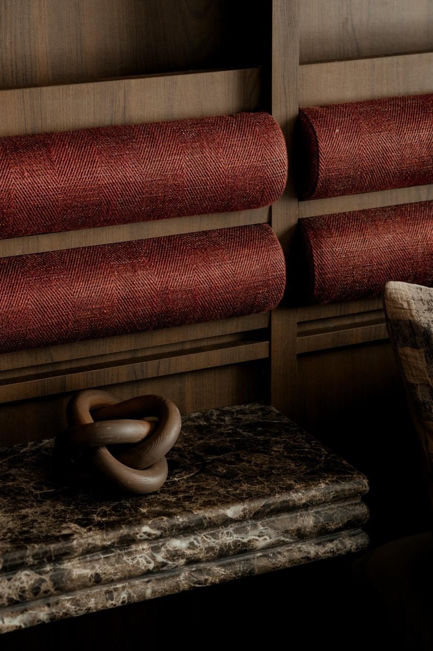 A close detail of the crimson rolled bolster cushions set into the wood-framed headboard panels, with a dark veined marble shelf and a wooden knot sculpture below