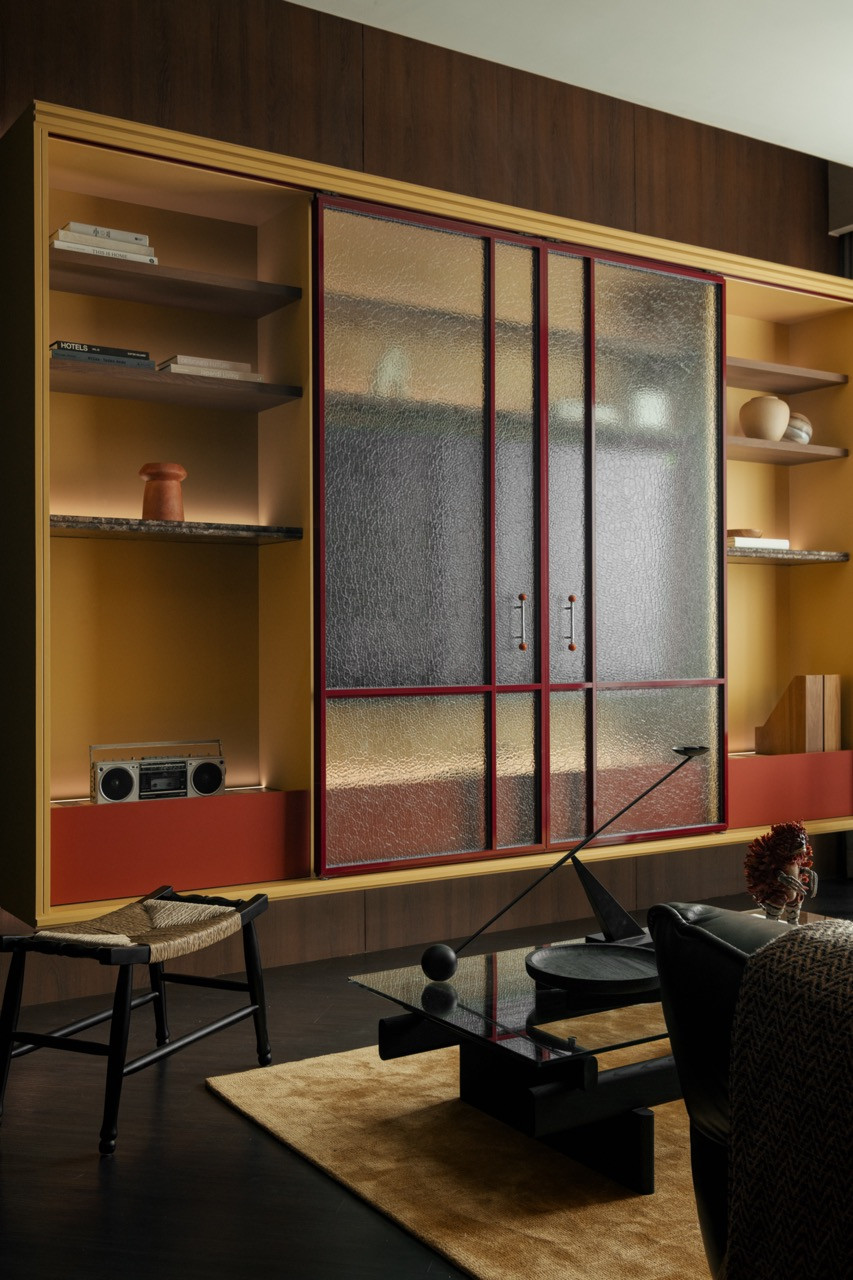 The living room display cabinet closed, its reeded glass panels in crimson frames flanking the central shelving, with a woven stool and black coffee table in the foreground