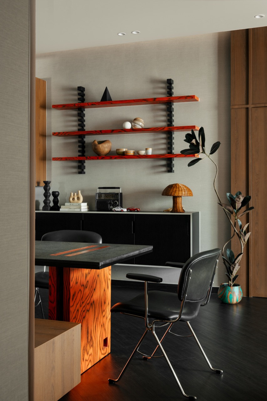 The dining area viewed from the living room, with the red-orange figured wood table base and open shelving beyond, beside a rubber plant in a turquoise ceramic pot
