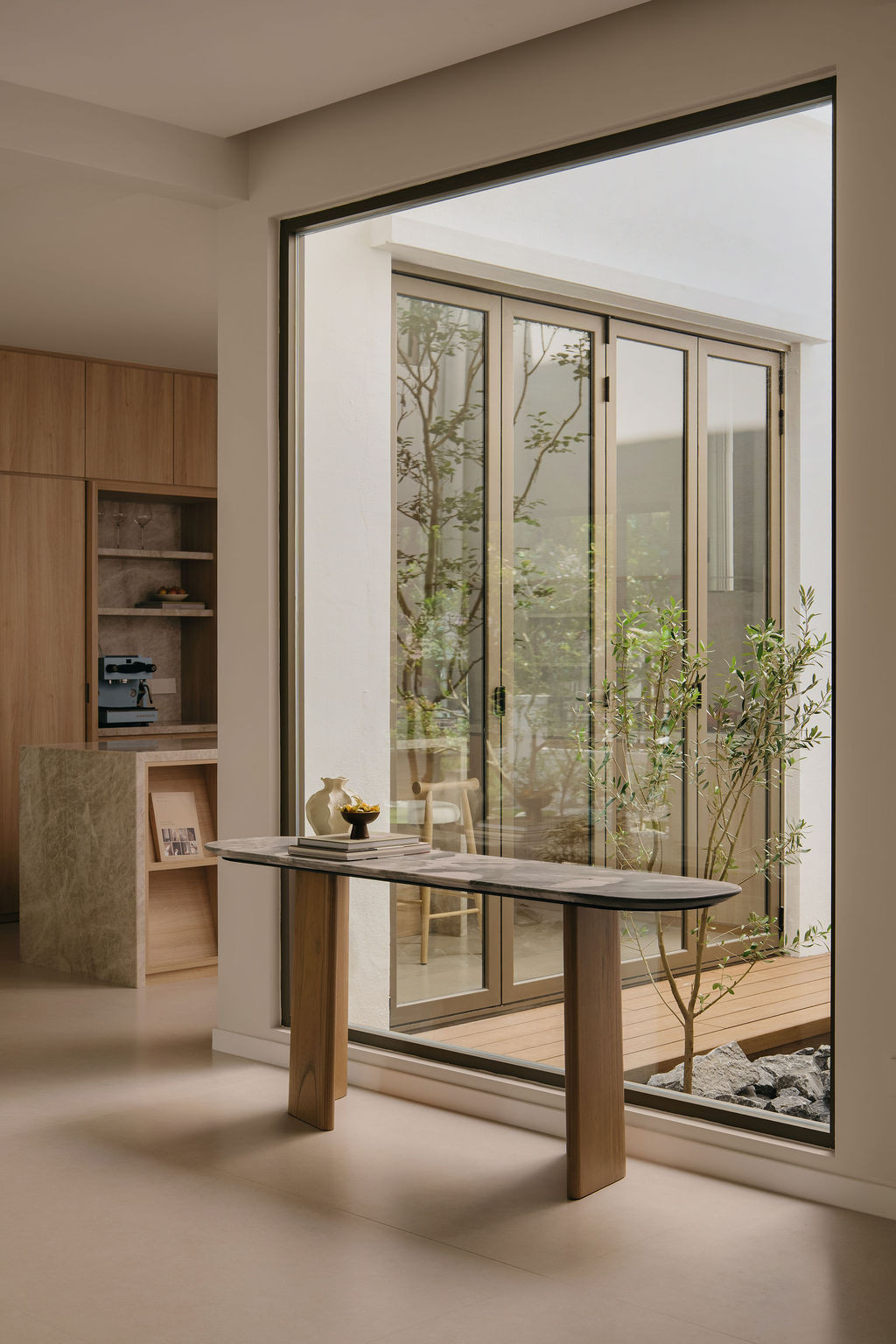 The Nicchris entrance console, stone-topped and styled with the homeowner’s decorative objects, is positioned in front of the picture window that frames the indoor courtyard and its Sputnik planting