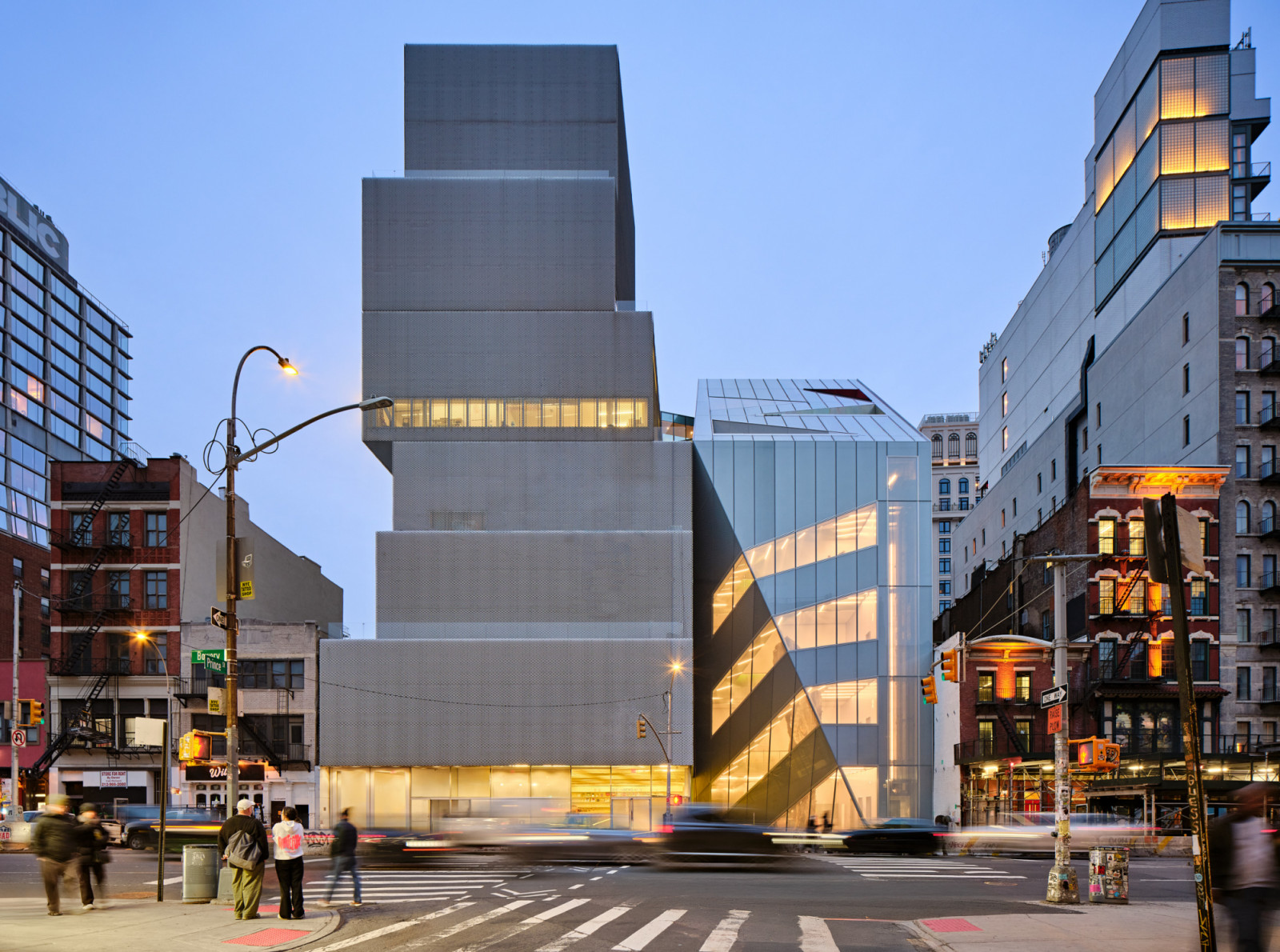 The expanded New Museum at dusk, viewed from the Bowery and Prince Street intersection. The OMA-designed addition’s angular glass facade meets the original SANAA building, whose stacked metal-mesh volumes rise to the left (Photo: New Museum)
