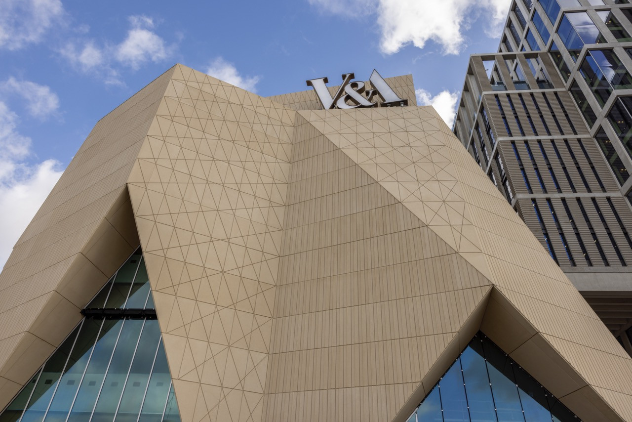 The faceted precast concrete facade of the V&A East Museum, its triangulated surface geometry catching afternoon light against a partly cloudy sky (Photo: V&A East)