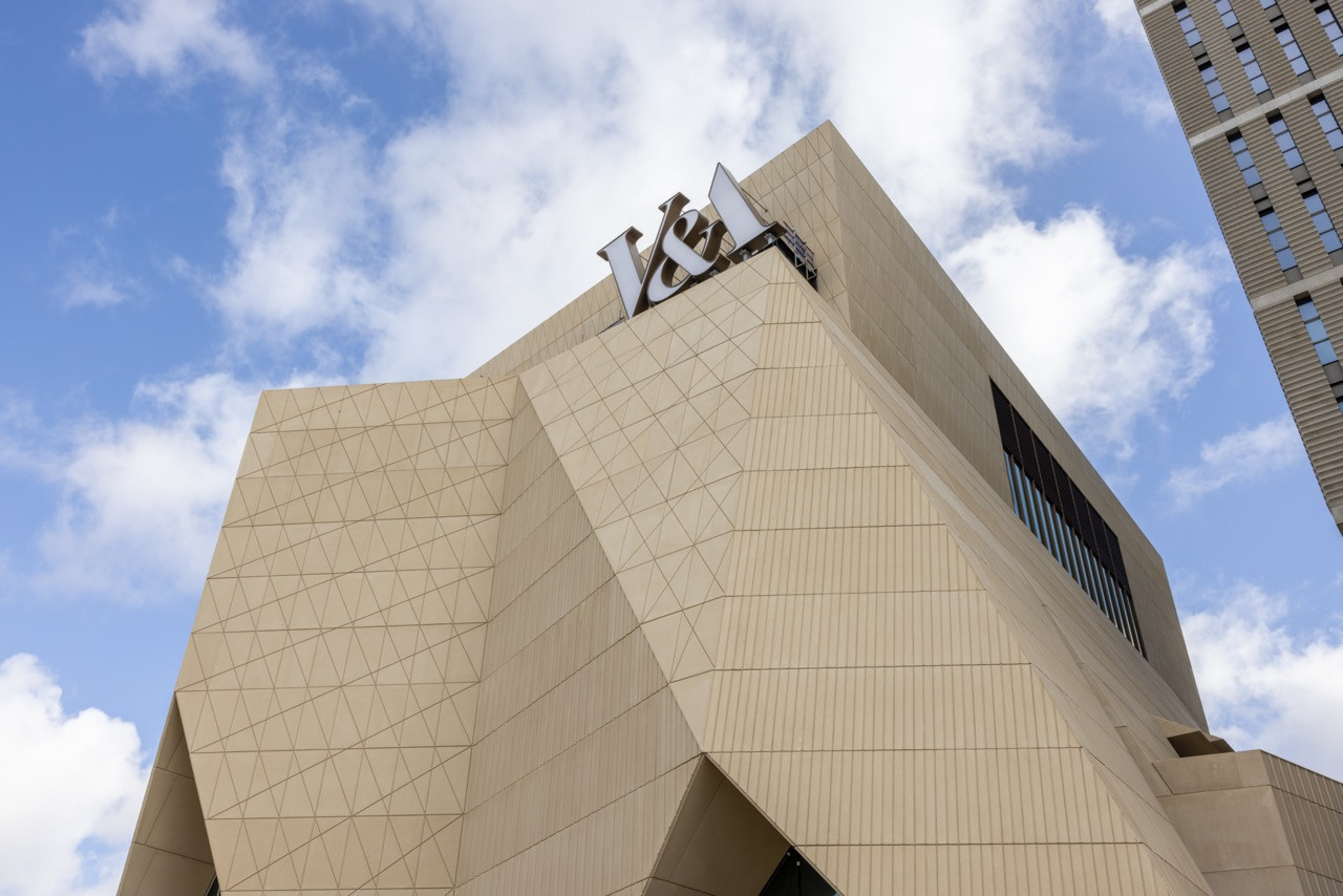 A close-up of the V&A East Museum’s upper facade, designed by O’Donnell + Tuomey, showing the scored and profiled precast concrete panels and the V&A lettering at the building’s crown (Photo: V&A East)
