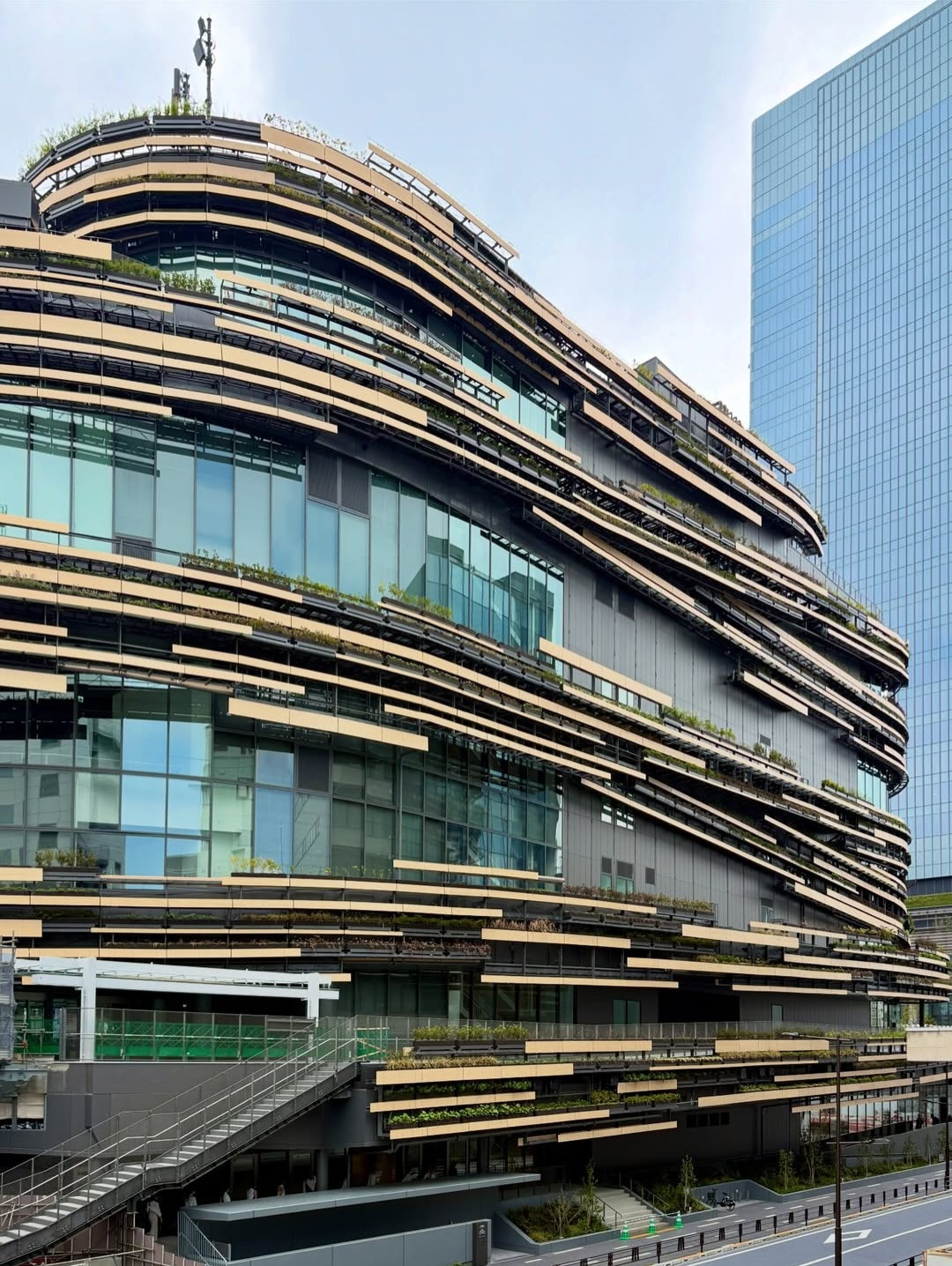 MoN Takanawa is seen from street level, its layered facade of warm timber panels and planted terraces contrasting with the glass towers of the surrounding development. The museum opened on 28 March 2026 as part of the Takanawa Gateway City cultural quarter. (Photo: Instagram/@281archdesign)