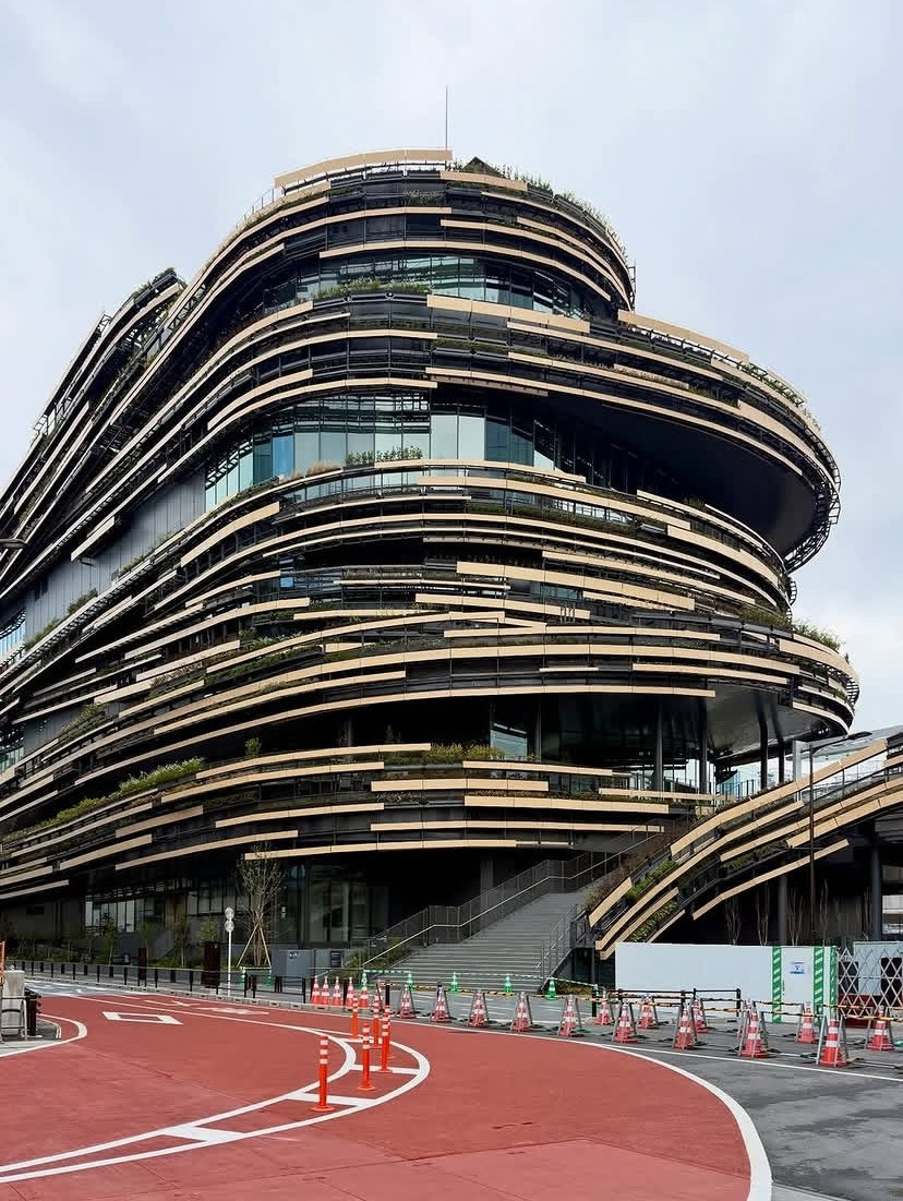 The spiral timber-clad form of MoN Takanawa: The Museum of Narratives, designed by Kengo Kuma & Associates, rising above the Takanawa Gateway City development in Minato, Tokyo. Terraced greenery runs between the building’s horizontal banding to the rooftop garden above (Photo: Instagram/@281archdesign)