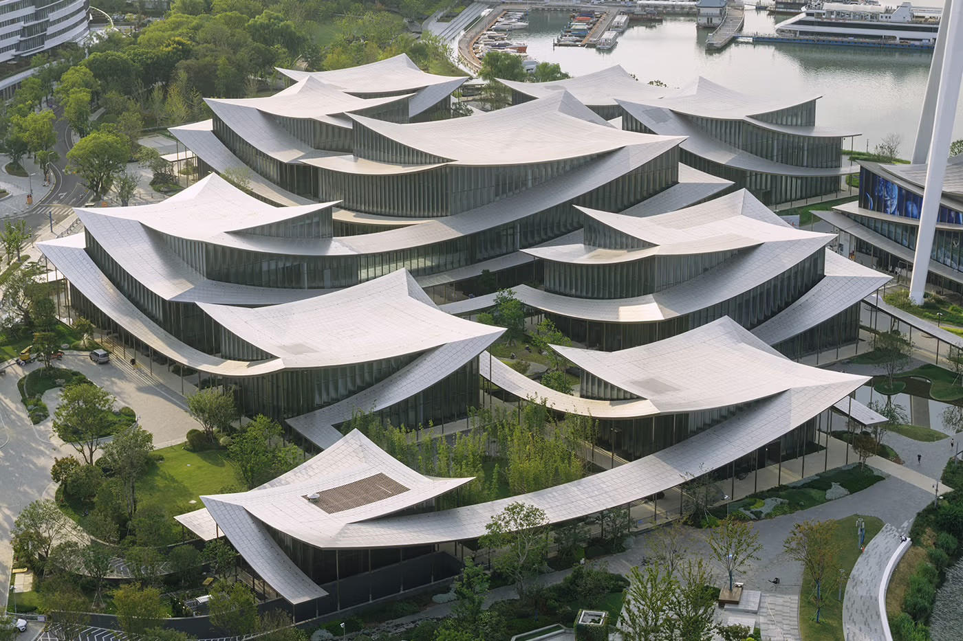 An aerial view of the full Suzhou Museum of Contemporary Art complex by day, showing the twelve interconnected pavilions and their continuous stainless steel roof across the Jinji Lake waterfront (Photo: BIG)