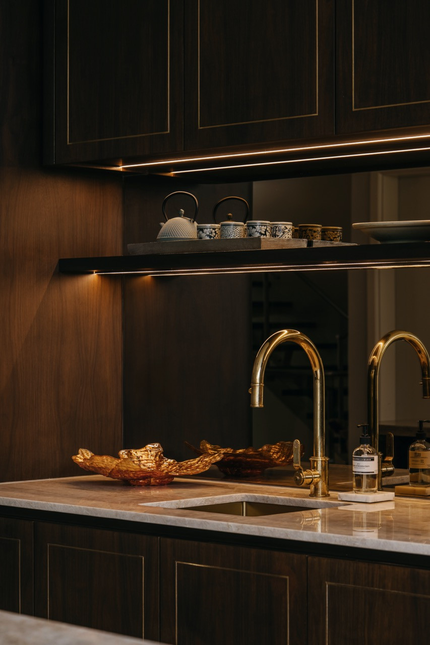 A detail of the wet kitchen counter with brass tap, mirrored splashback, under-shelf lighting, and a blue-and-white tea service on the open shelf above