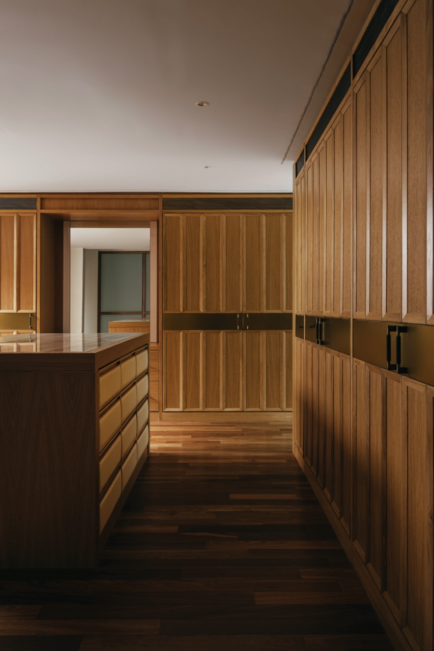 The wet kitchen, viewed from behind the island, with floor-to-ceiling oak cabinetry and brass inlay bands extending toward the brighter dry kitchen beyond