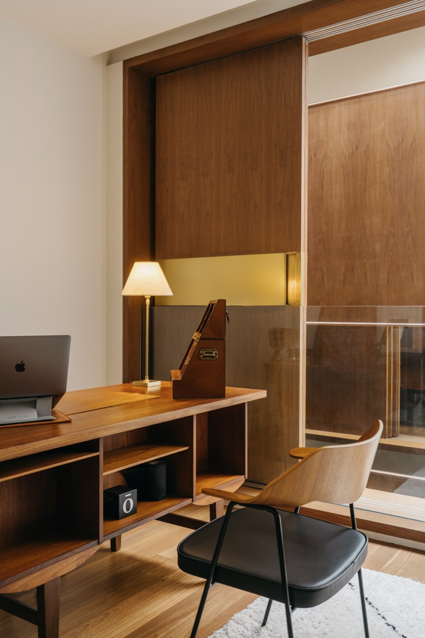 A home study with a walnut desk and brass table lamp, set against a walnut sliding panel that screens the balcony beyond