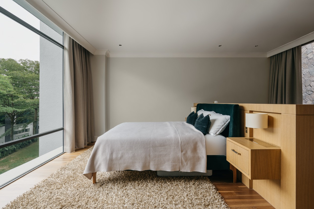 A guest bedroom with full-height glazing overlooking the tree canopy, with a teal upholstered headboard set against integrated oak bedside joinery