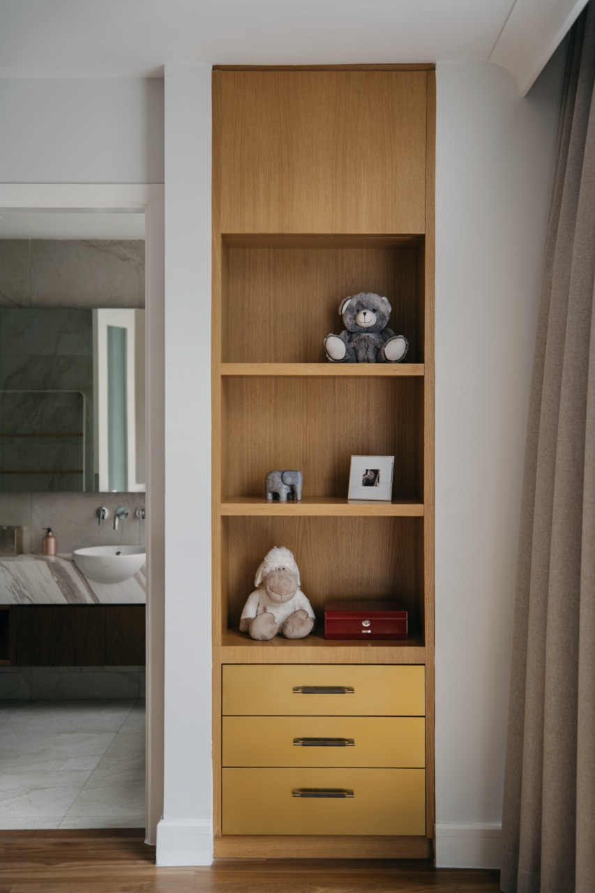 Oak open shelving with mustard-yellow drawers in a guest bedroom, with a glimpse of the marble-clad ensuite bathroom beyond
