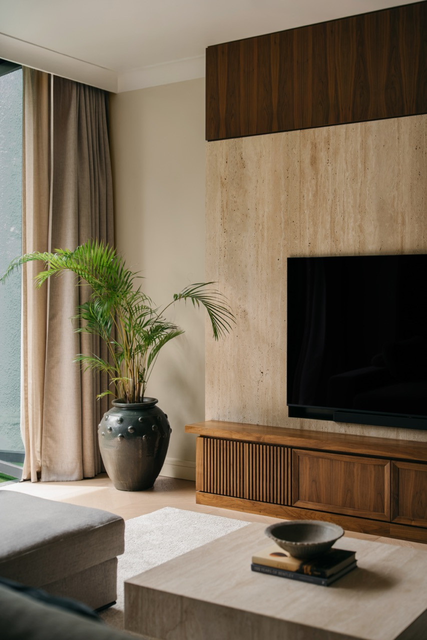 A detail of the living room’s travertine TV wall with walnut panel above, a slatted timber media cabinet below, and a large ceramic urn planted with an areca palm