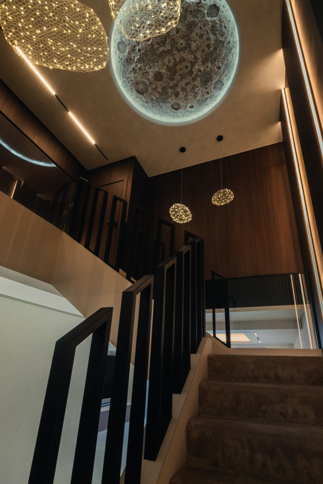 A view of the staircase, where sculptural black balusters frame the ascent while luminous ceiling and pendant lamps cast a glow across the double-height volume