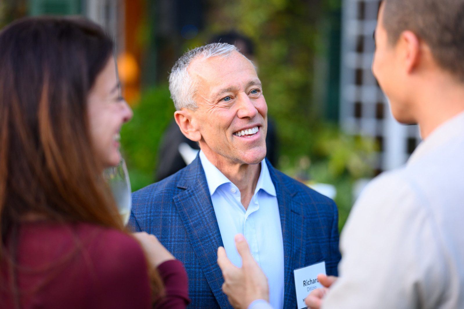Richard Ditizio, CEO of the Milken Institute, engaging in an animated conversation with guests during a reception at the Four Seasons Hong Kong. He is smiling warmly, illustrating the Institute's role as a "global matchmaker" in a sophisticated, social setting