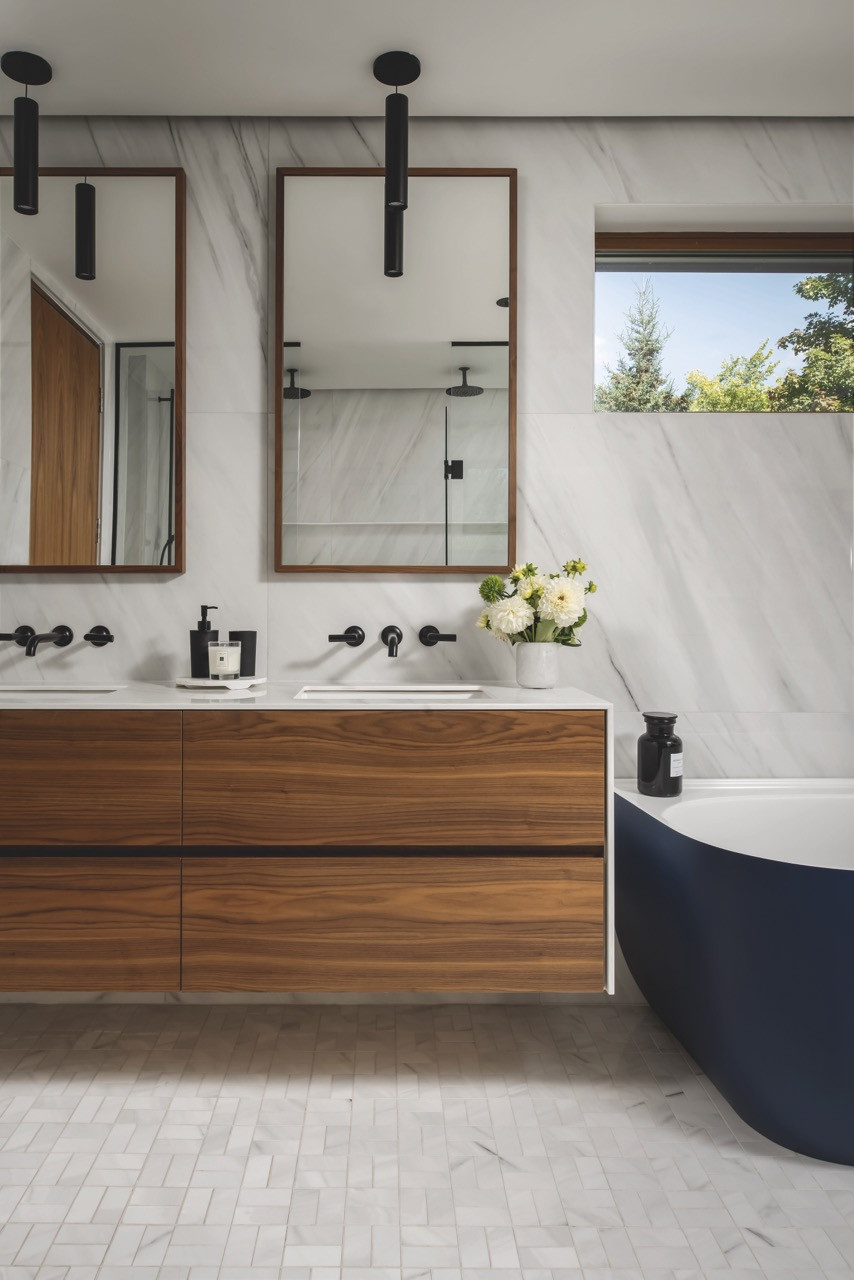 The primary ensuite is furnished with a walnut vanity, white porcelain-slab walls, black fixtures and a freestanding bathtub with views towards the lake