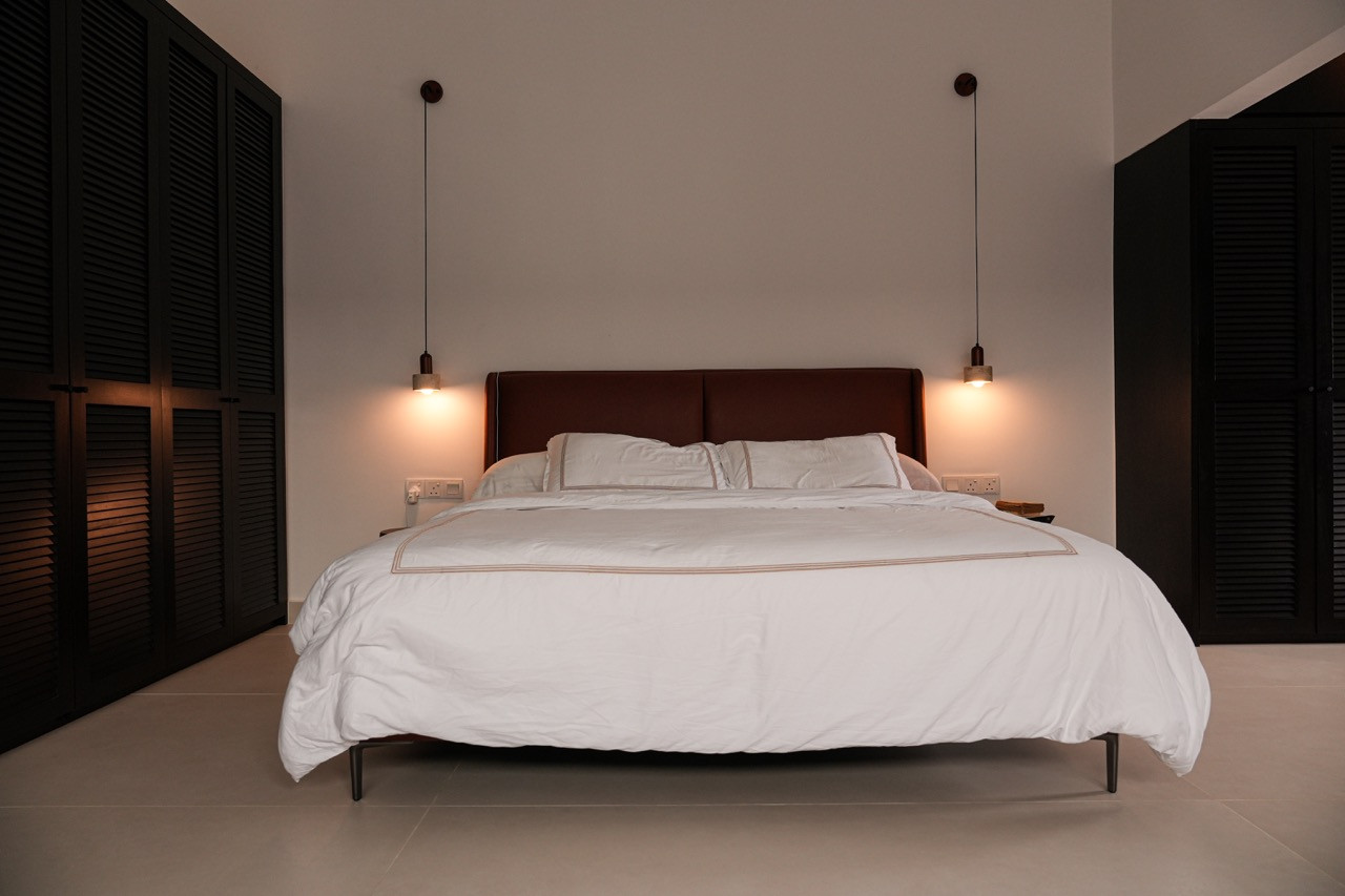 The master bedroom with a leather-upholstered bed, white linen, and paired pendant lights, flanked on both sides by dark, solid wood louvred wardrobe doors