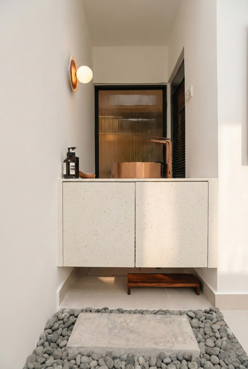 A bathroom vanity with a wall-mounted terrazzo cabinet, copper vessel sink, copper tap, and reeded glass panel, with river pebbles and a stepping stone on the floor