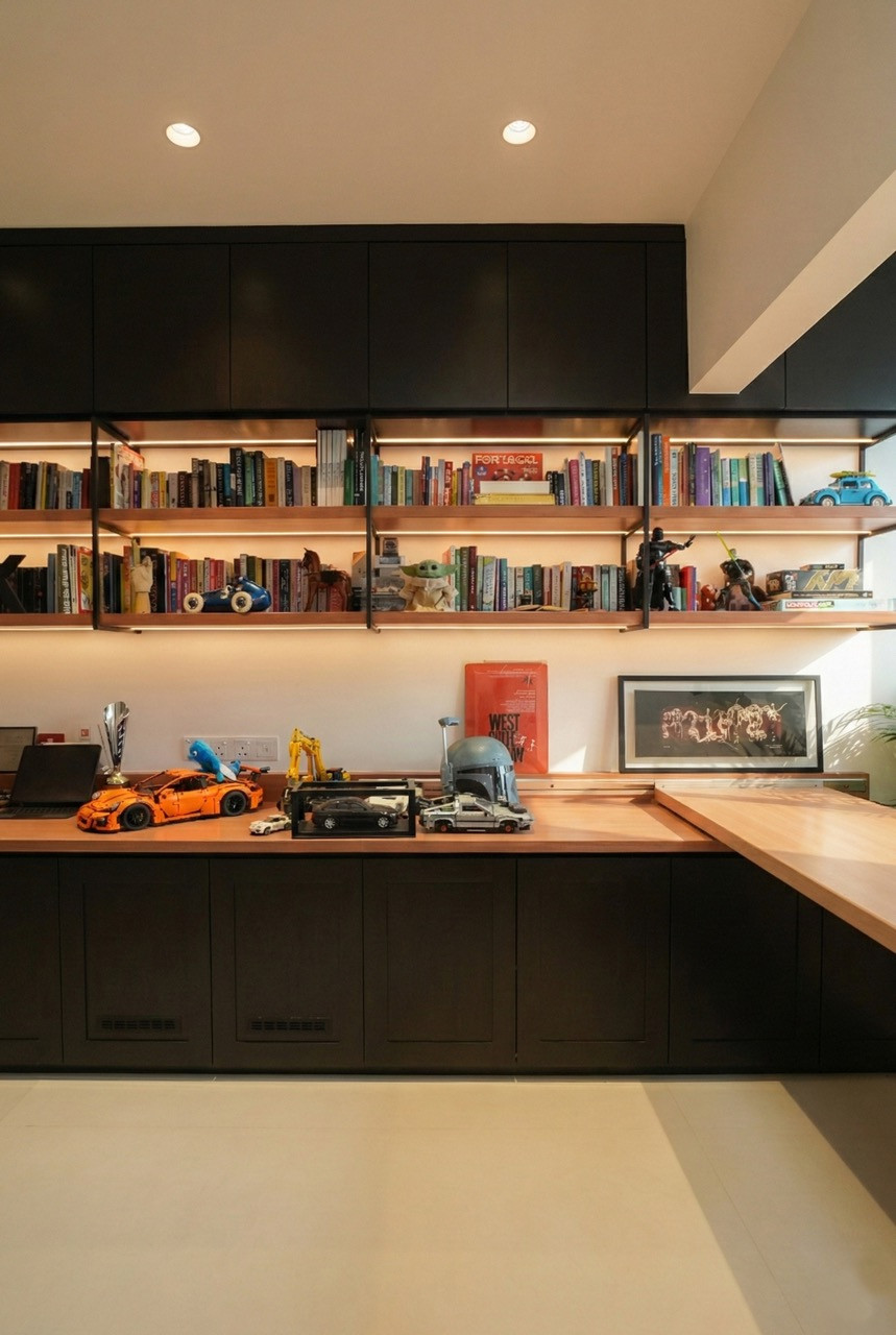 The study with dark wood cabinetry, backlit open shelving displaying books, scale car models, a Boba Fett helmet, and framed prints, with a warm timber desktop running the length of the wall