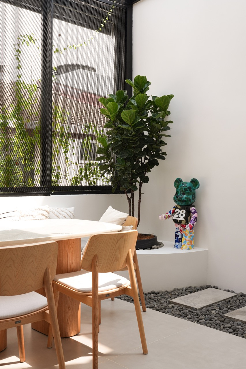 The dining area corner with a round marble table, timber chairs, a fiddle-leaf fig planted in a pebble-filled ledge, and a Bearbrick figure on the bench beside the perforated steel screening