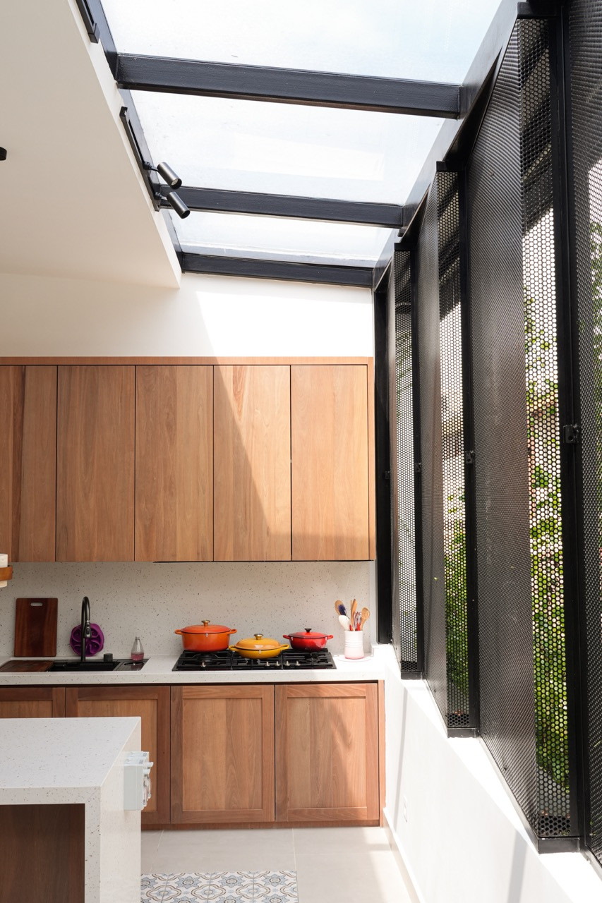 The kitchen showing solid wood upper and lower cabinetry, a terrazzo splashback, a gas hob, a glass roof, and open perforated steel louvre panels at the rear with climbing plants visible outside