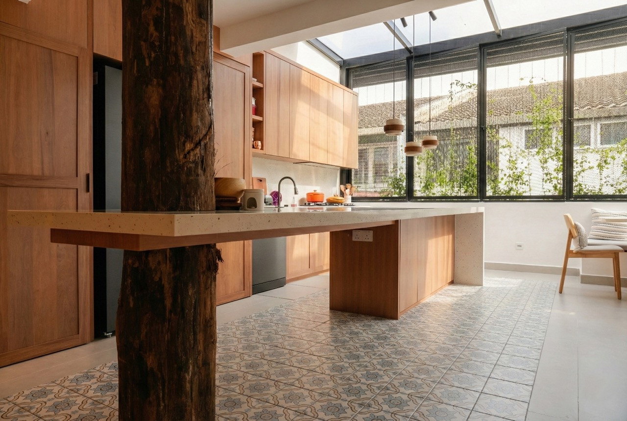 The kitchen with solid wood cabinetry, a terrazzo-topped island, encaustic patterned floor tiles, and a glass roof extension, with a raw timber post in the foreground and perforated steel rear screening with climbing plants beyond