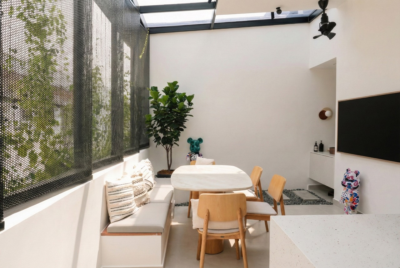 The rear dining area under a glass roof with perforated steel screening, a marble-topped table, timber chairs, built-in banquette seating, a fiddle-leaf fig, and a Bearbrick figure displayed on a ledge