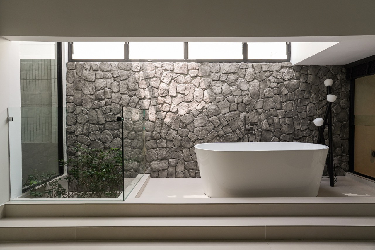 The Premtex freestanding bathtub set on a raised platform against the rough stone feature wall, with a glass shower enclosure to the left and a multi-globe floor lamp to the right