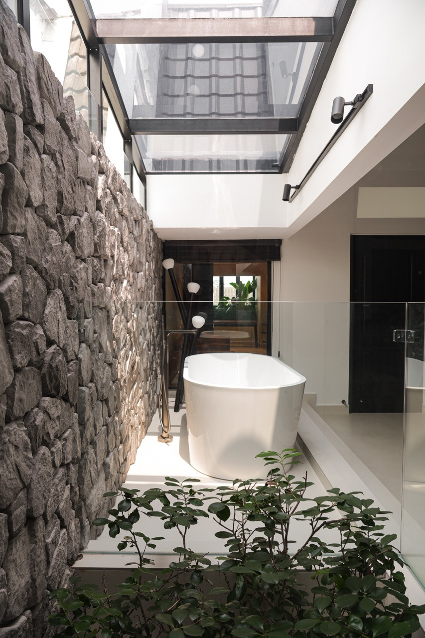 The master bathroom freestanding white bathtub positioned beneath a glass roof beside a rough stone feature wall, with small plants in the foreground and a view through to the bedroom beyond