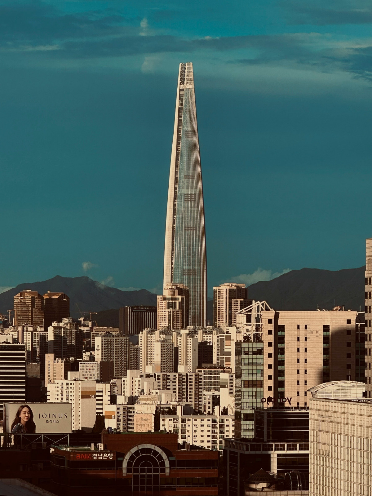 Lotte World Tower seen against the Seoul skyline and the mountains of Bukhansan beyond, its rounded profile contrasting with the denser residential and commercial fabric of the surrounding Jamsil district (Photo: Unsplash/Yeon Lee)