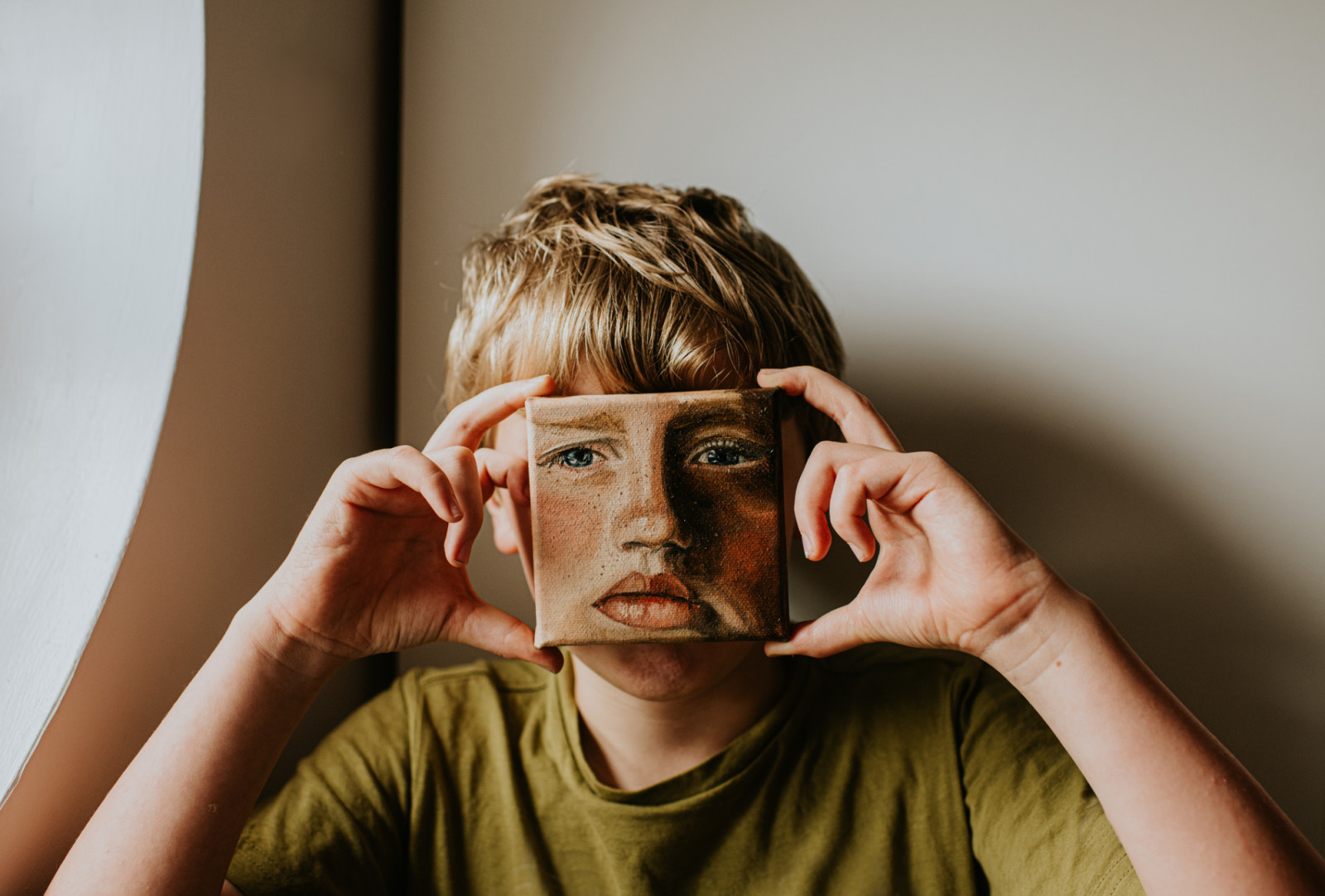 A young lad covers his face with a small oil painting of himself. The expression on the painting is serious. Conceptual with space for copy.