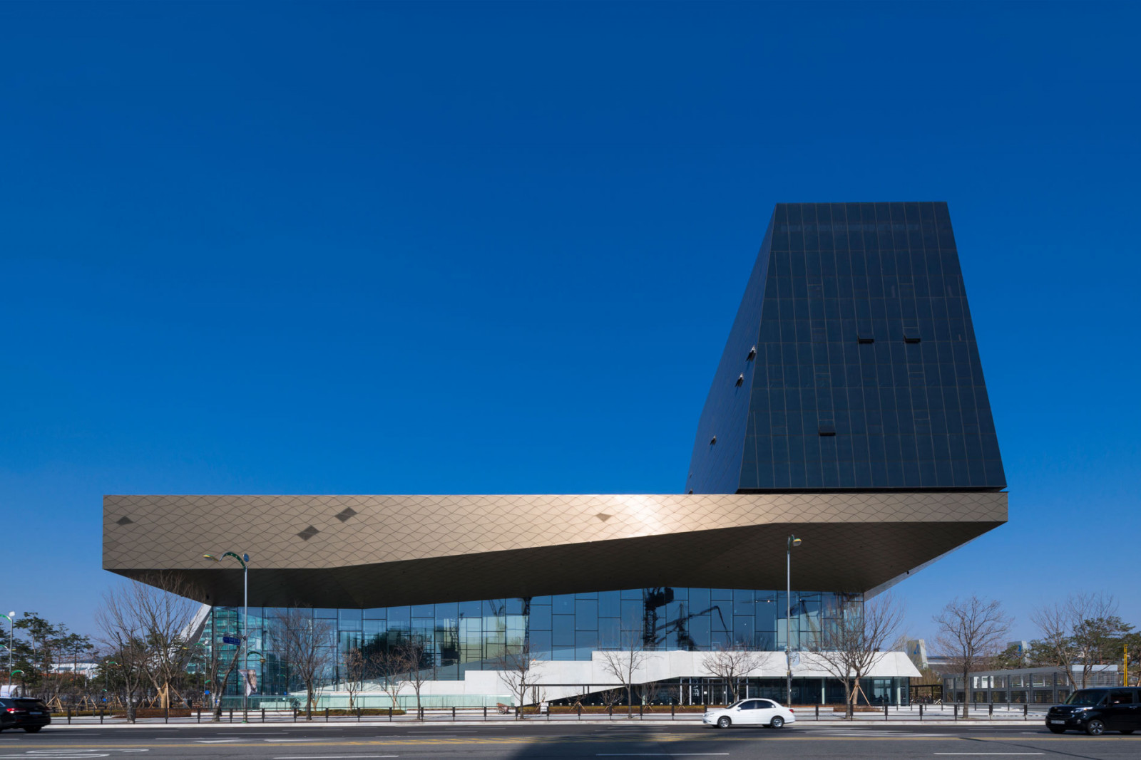 The street-facing façade of Hyundai Motorstudio Goyang, designed by Delugan Meissl Associated Architects and completed in 2017, shows the cantilevered “shaped sky” roof — clad in bronze-toned aluminium panels — extending over a fully glazed ground-level volume (Photo: Instagram/DMAA)