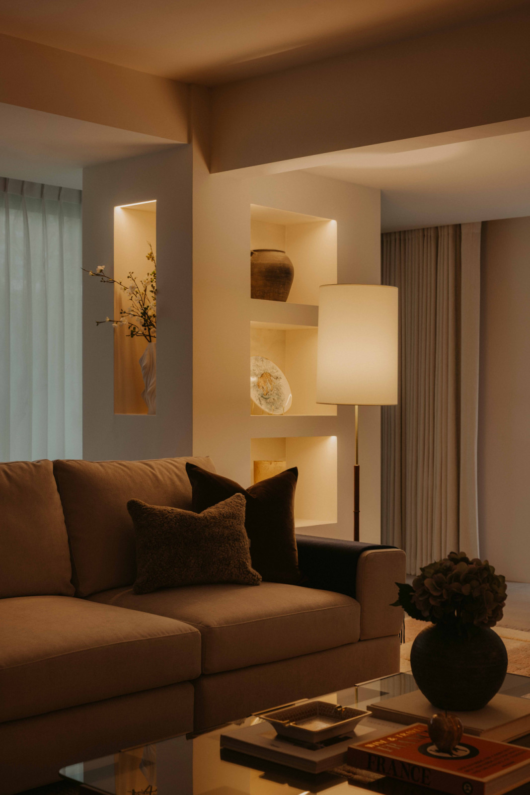 Built-in wall niches and a floor lamp bring warmth and depth to the living area, where a wall sconce from Luke Lamp Co reads like an artwork